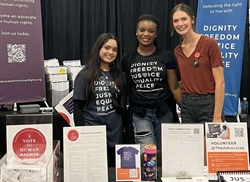 [ai] Three young women at a booth promoting human rights, wearing black shirts with the text "DIGNITY FREEDOM JUSTICE EQUALITY PEACE." The table has printed materials and items for awareness.
